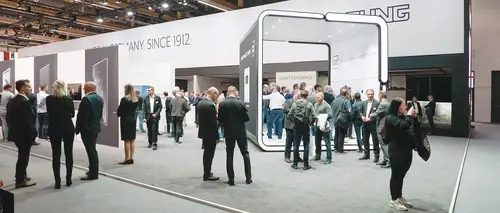 Messestand auf einer Fachmesse. Zahlreiche Besucher stehen in Gruppen, unterhalten sich oder erkunden die ausgestellten Produkte. Der Stand hat ein modernes Design mit klaren Linien und einer offenen Struktur.