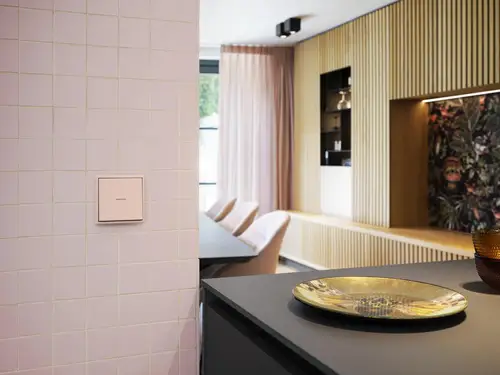Hellrosa gekachelte Wand mit einem JUNG HOME Taster, schwarzer Tisch mit goldener Schale im Vordergrund und Sitzmöbel mit Regal im Hintergrund.