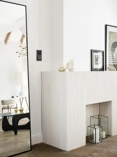 Ein modernes Interieur mit einem großen Spiegel, der eine Tischlampe und Holzbalken widerspiegelt. Neben dem Spiegel befindet sich ein JUNG HOME Raumthermostat und ein JUNG HOME Taster einfach in einer zweifachen Kombination an einer weißen Wand