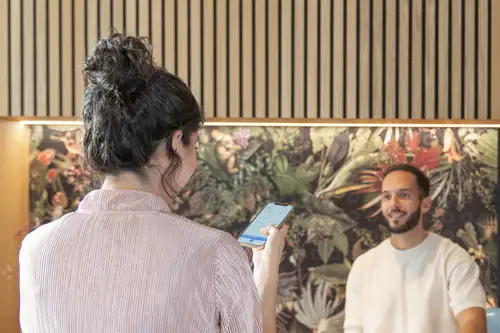 Frau betrachtet Smartphone mit der JUNG HOME App, während Mann ihr gegenübersteht; auffällige botanische Wanddekoration im Hintergrund.