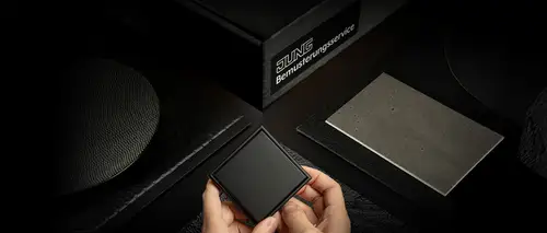 Schalter im Programm LS 990 in Graphitschwarz matt. Ambienteaufnahme mit Händen die den Schalter festhalten und verschiedenen Materialien im Umfeld.