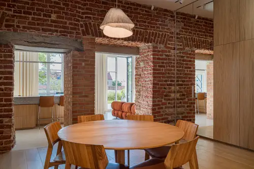 Ambient shot in a private residence in Vilnius. Section of a room with a round dining table and six chairs in the foreground, all made of wood. In the background there is a brick passageway and a mirrored built-in wardrobe on the right.