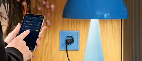 JUNG HOME Steckdose im Programm Les Couleurs® Le Corbusier®. Ambienteaufnahme in der eine Hand die Smart Visu App bedient.