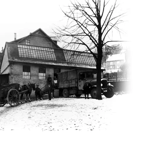 Black and white photograph of a building with a pitched roof and large windows. A lorry and a horse-drawn cart are parked in front of the building. Two men are working on the horse-drawn cart while other people are standing by the lorry. Snow-covered houses and trees can be seen in the background.