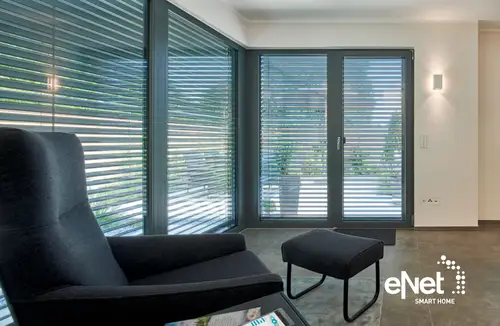 Logotipo de eNet SMART HOME en un moderno salón con grandes ventanales y persianas eléctricas. Sillón y taburete negros en primer plano.