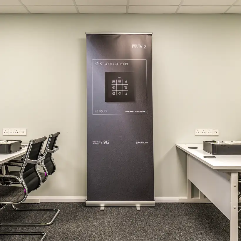 JUNG training centre in London, ambience shot in the training room with JUNG products as teaching material. The focus is on a dark blue stand with a KNX room controller in the LS Touch range in black.