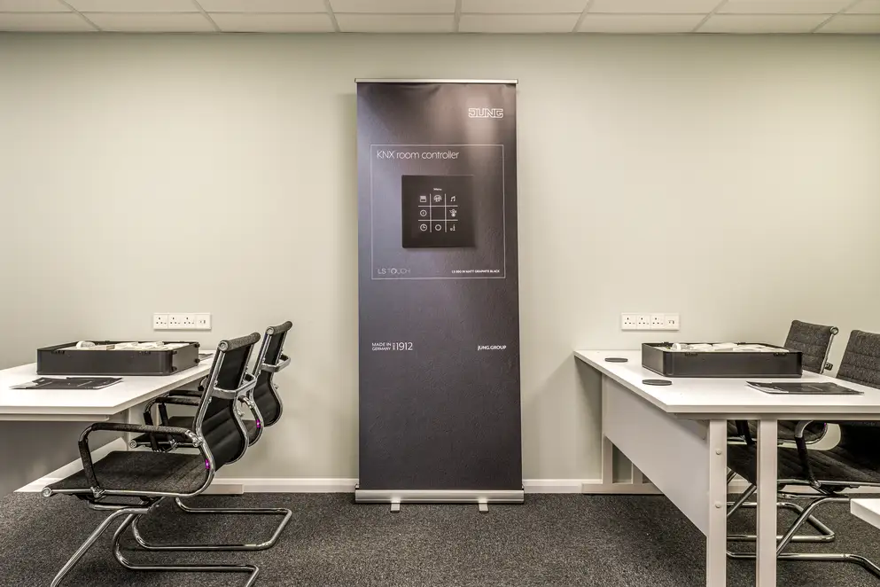 JUNG training centre in London, ambience shot in the training room with JUNG products as teaching material. The focus is on a dark blue stand with a KNX room controller in the LS Touch range in black.