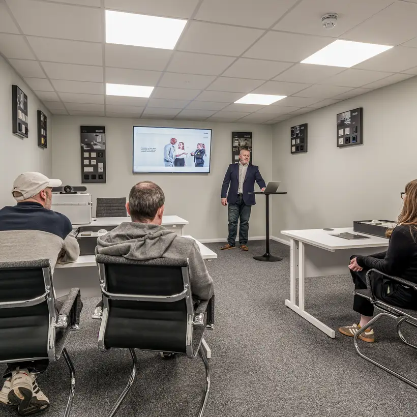 Trainees receive instruction in the training room of the JUNG training centre in London. The trainee standing at a lectern is at the front at the back of the room and the three trainees are sitting with their backs to the photographer.