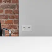 Frontalansicht von 2 LS 990 Steckdosen in Alpinweiß an einer weißen Wand. Neben den Steckdosen befinden sich eine Backsteinwand und ein Bürostuhl