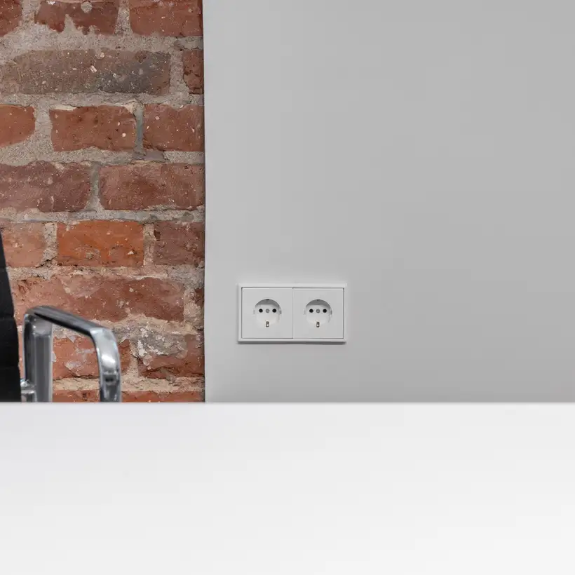 Frontalansicht von 2 LS 990 Steckdosen in Alpinweiß an einer weißen Wand. Neben den Steckdosen befinden sich eine Backsteinwand und ein Bürostuhl