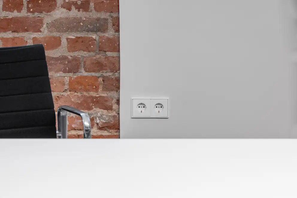 Frontalansicht von 2 LS 990 Steckdosen in Alpinweiß an einer weißen Wand. Neben den Steckdosen befinden sich eine Backsteinwand und ein Bürostuhl