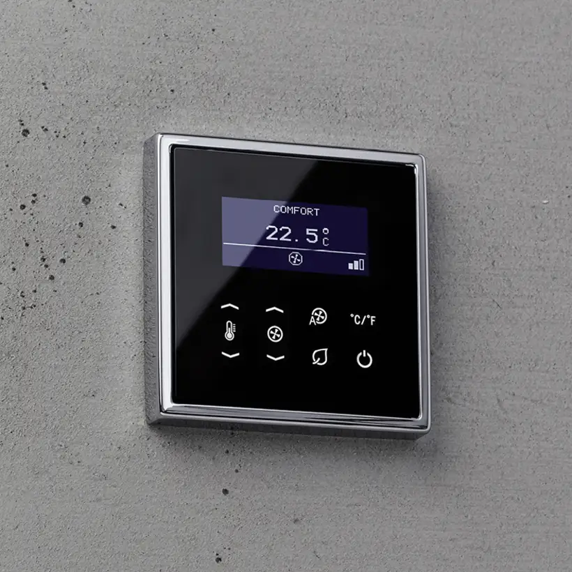 Raumtemperaturregler FanCoil im Programm LS 990 mit einem Glanzchromrahmen und schwarzem Display. Ambienteaufnahme auf einer Betonwand in leichter Perspektive.