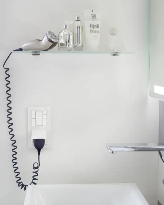 Taster F 50 für KNX und eNet SMART HOME und Steckdose im Programm A Creation aus Glas in Alpinweiß. Frontal Fotografiert, ein elektrischer Rasierapparat steckt in der Steckdose und liegt auf einem Glasregal, neben Flacons, oberhalb des Tasters.