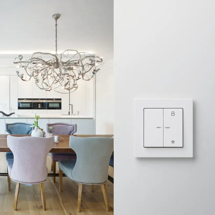 KNX Taster als Serienwippe im Programm A Creation aus Kunststoff in Alpinweiß, frontal aufgenommen, in einem Durchgang. Linksseitig befindet sich im Hintergrund ein Essbereich mit weißer Küchenzeile.