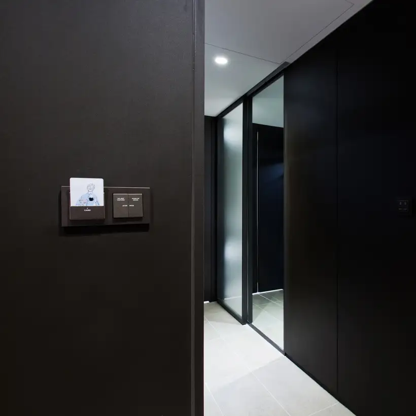 Hotelkartenhalter und KNX Taster als Serienwippe im Programm A Creation aus Kunststoff, Ambienteaufnahme in leichter Perspektive auf einer dunklen Holzwand. Küchenzeile und Flur des Hotelzimmers im Ausschnitt.