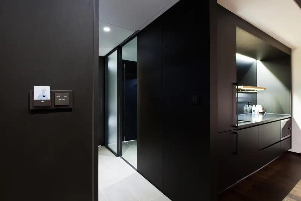 Hotelkartenhalter und KNX Taster als Serienwippe im Programm A Creation aus Kunststoff, Ambienteaufnahme in leichter Perspektive auf einer dunklen Holzwand. Küchenzeile und Flur des Hotelzimmers im Ausschnitt.