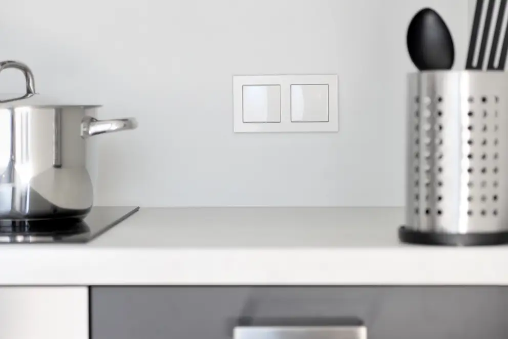 Taster im Programm A Creation aus Kunststoff in Alpinweiß, frontal fotografiert mit geringer Tiefenschärfe, so dass die im Ausschnitt gezeigte Küchenzeile und die darauf stehenden Töpfe und Utensilien eine Unschärfe haben.