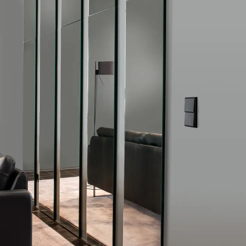 Taster im Programm LS 990 in Graphitschwarz matt, Ambienteaufnahme in leichter Perspektive, auf einer grauen Wand. Vordergründig steht ein Ledersofa mit Stehlampe, linksseitig hängen mehrere Spiegelpanele.