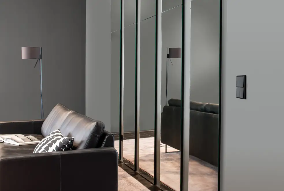 Taster im Programm LS 990 in Graphitschwarz matt, Ambienteaufnahme in leichter Perspektive, auf einer grauen Wand. Vordergründig steht ein Ledersofa mit Stehlampe, linksseitig hängen mehrere Spiegelpanele.