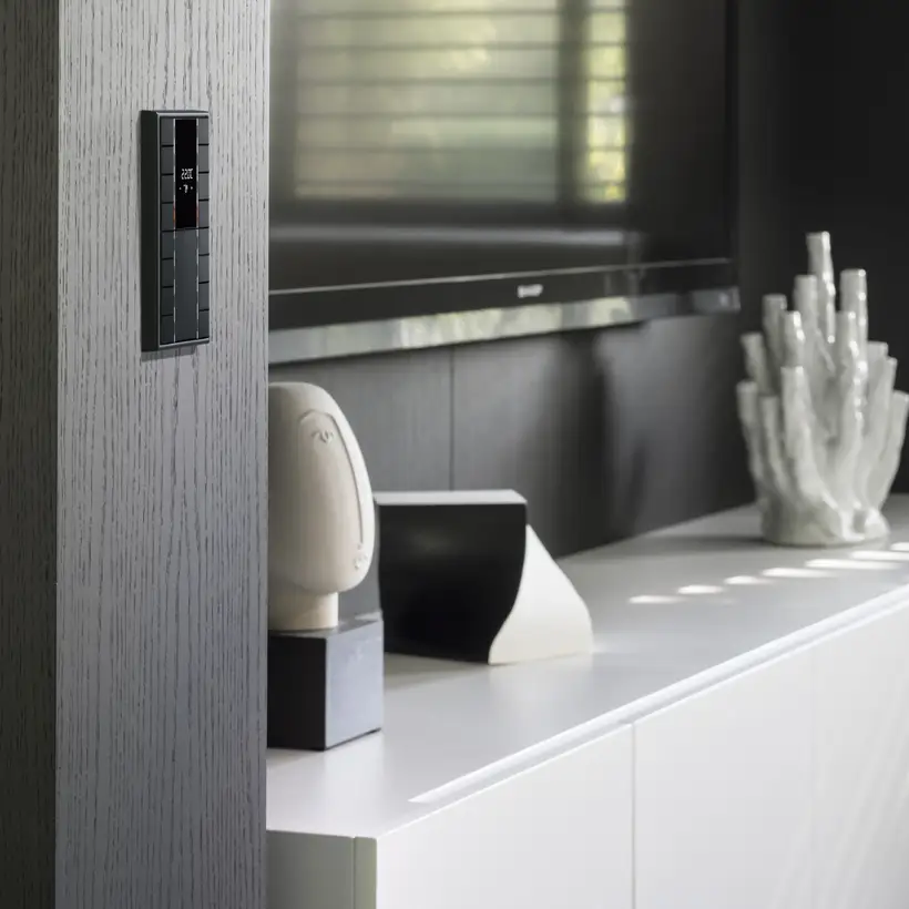 KNX F 50 4fach Taster im Programm LS 990 in Graphitschwarz matt, fotografiert in leichter Perspektive, auf einer in Holzvertäfelten Wand in anthrazit. In einer Niesche daben steht ein Lowboard in weiß mit Dekoelementen und darüber hängt ein TV Gerät.