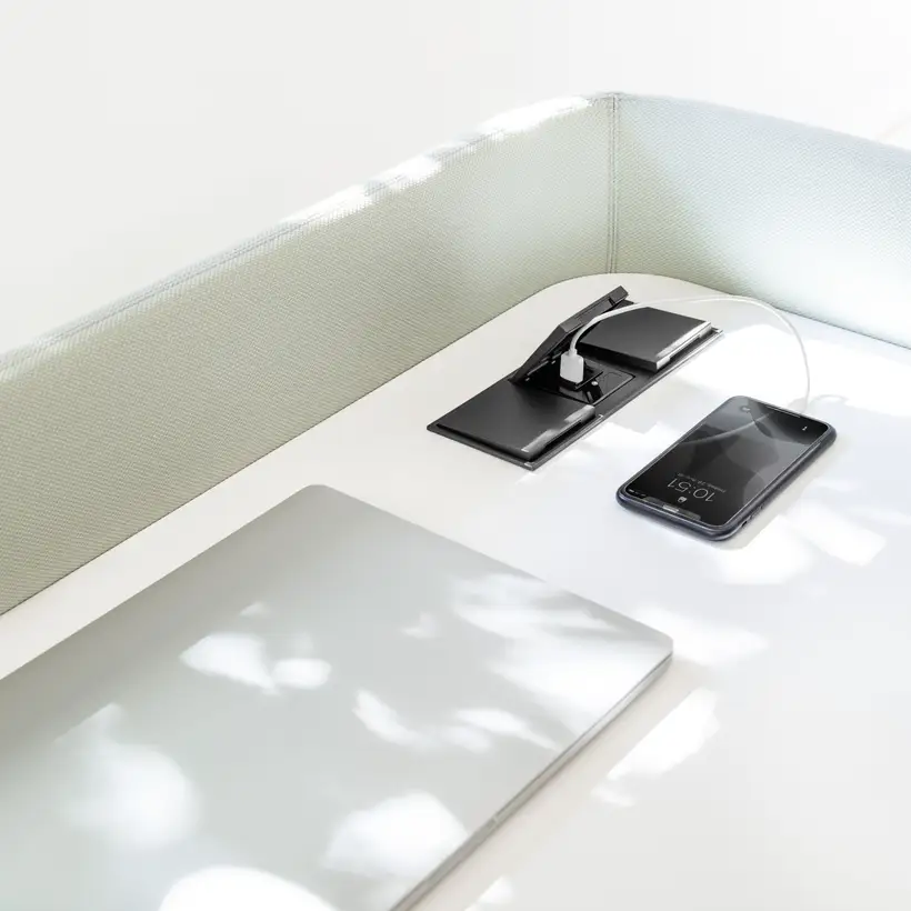 Raumausschnitt im COR Haus Nicola Rehage Ausstellungsraum, USB-Ladegeräte mit Anschlüssen für USB Typ A und Typ C integriert in einen weißen Tisch. Ein IPhone wird soeben geladen und ein geschlossenes MacBook liegt auf dem Tisch.