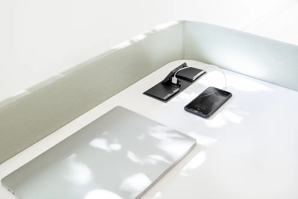 Raumausschnitt im COR Haus Nicola Rehage Ausstellungsraum, USB-Ladegeräte mit Anschlüssen für USB Typ A und Typ C integriert in einen weißen Tisch. Ein IPhone wird soeben geladen und ein geschlossenes MacBook liegt auf dem Tisch.