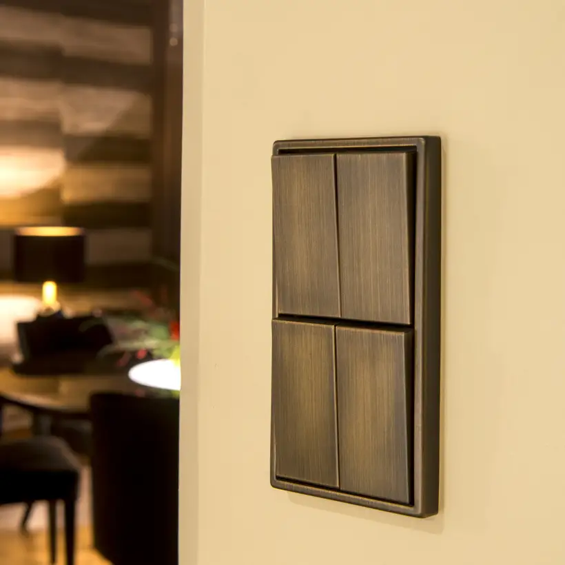 Serientaster im Programm LS 990 in Messing Antik, Close up in leichter Perspektive, auf einer Wand in Ecru im Hintergrund angeschnitten ein Tisch mit zwei Stühlen und eingeschalteter Tischleuchte.