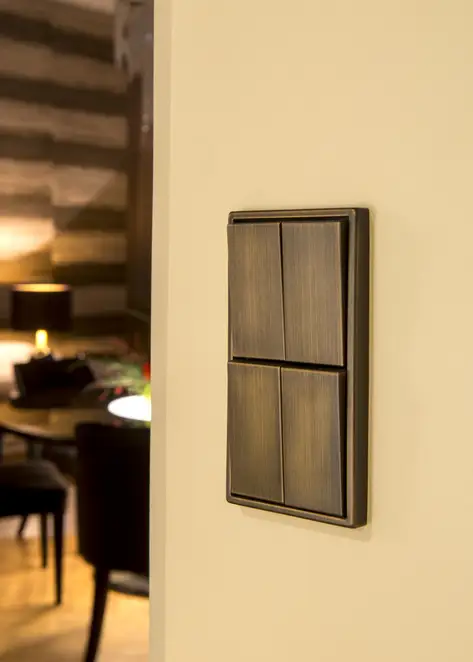 Serientaster im Programm LS 990 in Messing Antik, Close up in leichter Perspektive, auf einer Wand in Ecru im Hintergrund angeschnitten ein Tisch mit zwei Stühlen und eingeschalteter Tischleuchte.