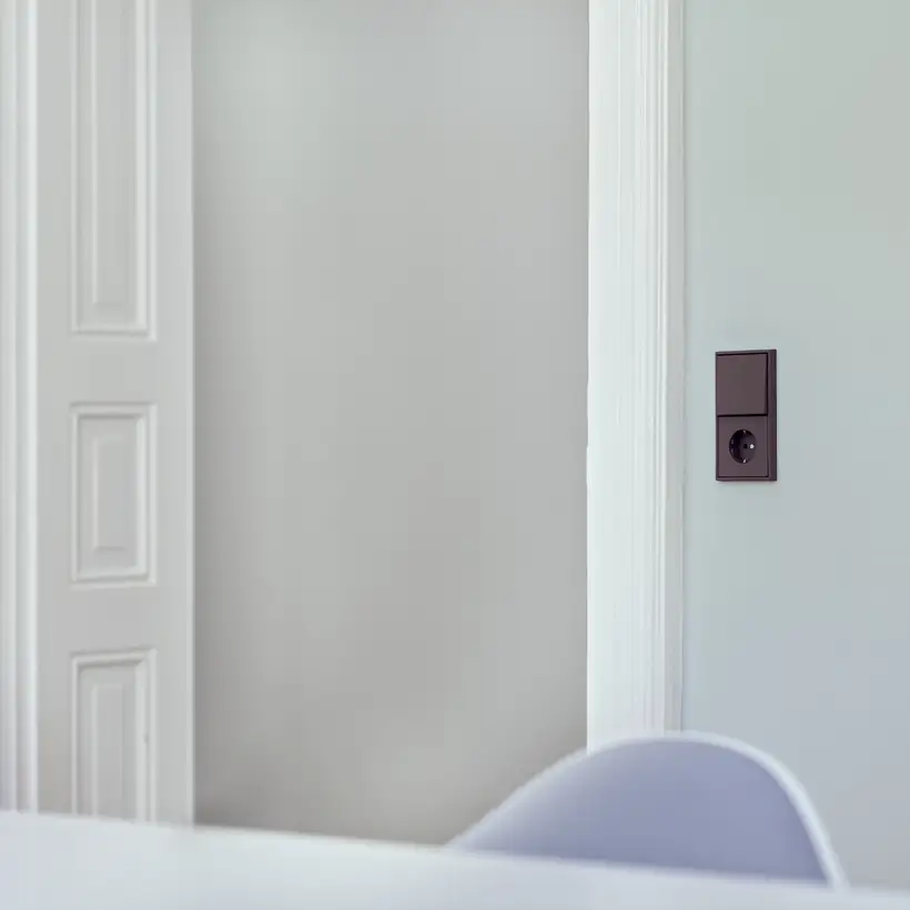 Ambienteaufnahme des LS 990 Taster und SCHUKO® Steckdose in Les Couleurs® Le Corbusiers universelles Farbsystem. Aufgenommen in leichter Perspektive, auf einer hellblauen Wand, neben Weiß lackierten Holztüren. Durch die geringe Tiefenschärfe sind Tisch un