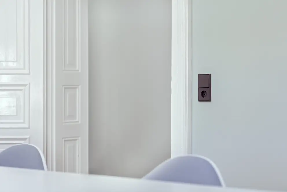 Ambienteaufnahme des LS 990 Taster und SCHUKO® Steckdose in Les Couleurs® Le Corbusiers universelles Farbsystem. Aufgenommen in leichter Perspektive, auf einer hellblauen Wand, neben Weiß lackierten Holztüren. Durch die geringe Tiefenschärfe sind Tisch un