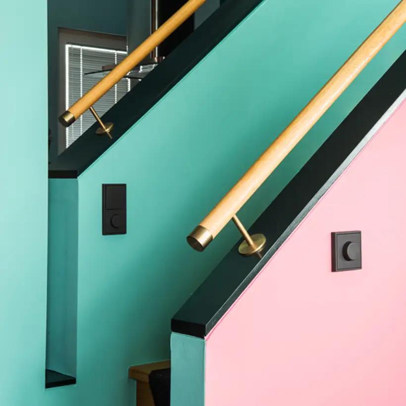 Der LS 990 Drehdimmer in Les Couleurs® Le Corbusier universellem Farbsystem, fotografiert in leichter Perspektive, an einem Pink gestrichenen Treppenaufgang mit schwarzen Akzenten und Holzhandlauf. 