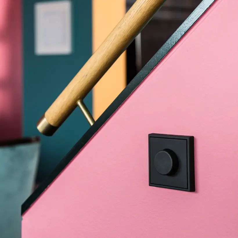 Der LS 990 Drehdimmer in Graphitschwarz matt, fotografiert in leichter Perspektive, an einem Pink gestrichenen Treppenaufgang mit schwarzen Akzenten und Holzhandlauf. 
