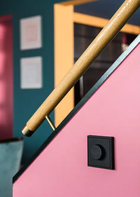 Der LS 990 Drehdimmer in Graphitschwarz matt, fotografiert in leichter Perspektive, an einem Pink gestrichenen Treppenaufgang mit schwarzen Akzenten und Holzhandlauf. 