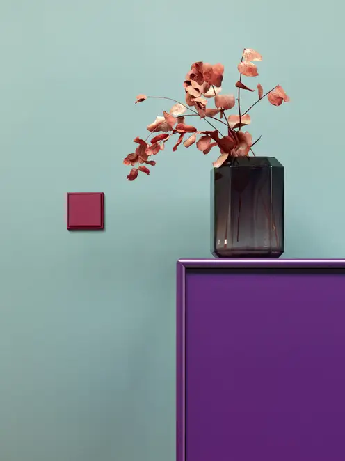 Ambienteaufnahme des LS 990 Taster in Les Couleurs® Le Corbusier in 32101 rouge rubia, frontal aufgenommen, auf einer petrolfarbenen Wand und neben einem Sideboad in Violett, auf welchem eine Vase mit rotem Eukalyptus steht.