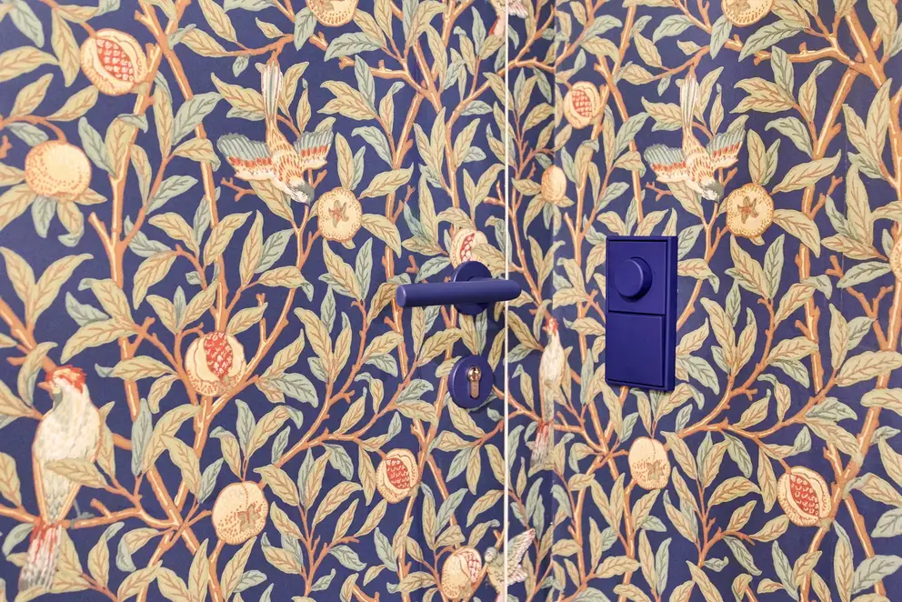 LS 990 Drehdimmer und Taster in Les Couleurs® Le Corbusier universellem Farbsystem, Ambienteaufnahme „Wohnen mit Morris“ Apartment in Stuttgart. Taster und Drehdimmer sind farblich passend zu dem Blau der floralen Tapete gewählt. Fotografiert in leichter 