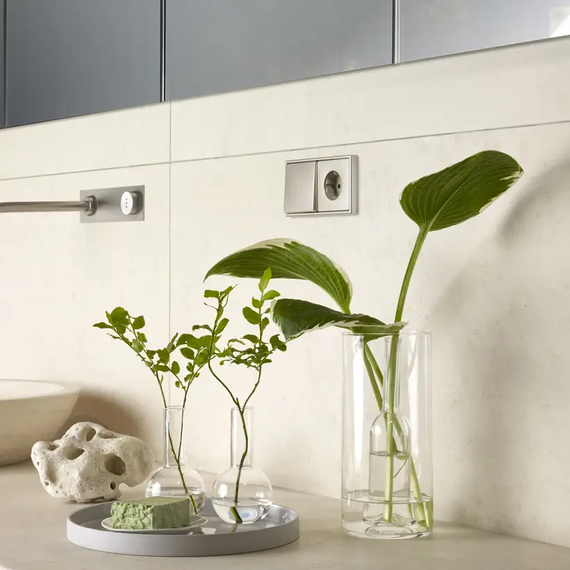 LS 990 SCHUKO® Steckdose und Taster aus Edelstahl, fotografiert in leichter Perspektive, über einer Steingut-Küchenzeile. Es stehen Pflanzenblätter in Vasen neben zwei runden Steingut-Waschbecken.