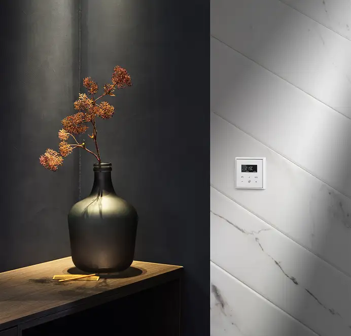 Raumthermostat im Programm LS 990 in Alpinweiß. Ambienteaufnahme vor einer stilvollen Marmor-Wand, mit moderner Vase. 