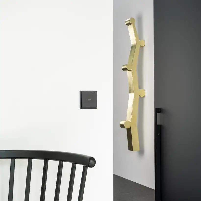 JUNG HOME Taster im Programm LS 990 in Graphitschwarz matt. Ambiente Aufnahme in einem modernen Flur mit goldenen Akzenten.