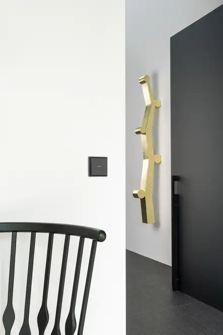 JUNG HOME Taster im Programm LS 990 in Graphitschwarz matt. Ambiente Aufnahme in einem modernen Flur mit goldenen Akzenten.